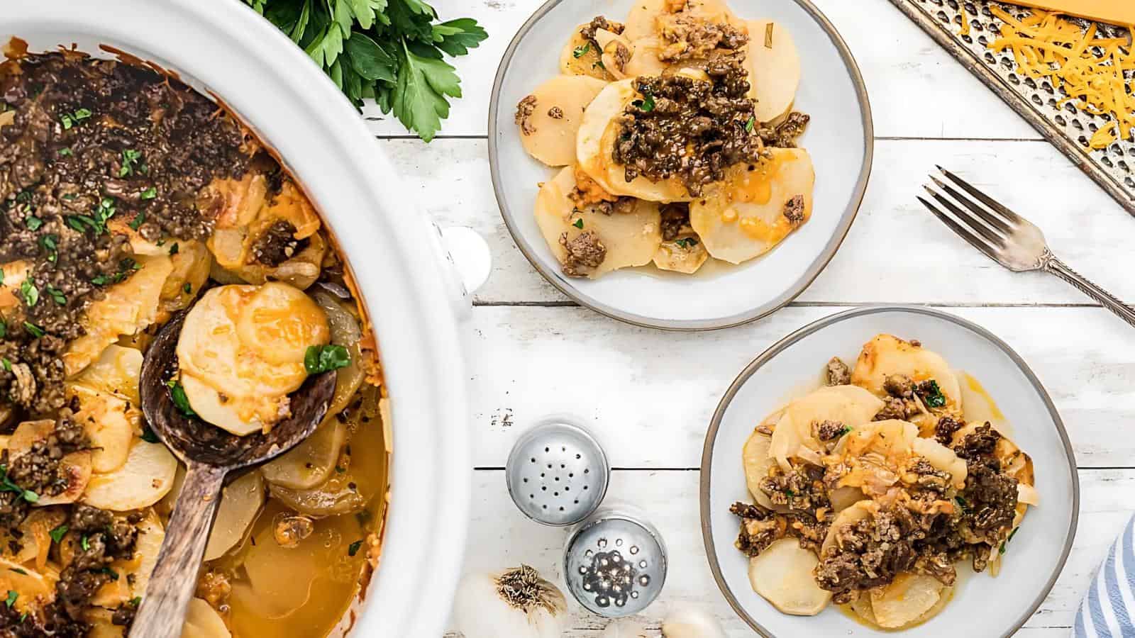 Top-down view of two plates and a serving dish with sliced potatoes and ground beef casserole, garnished with parsley, on a white wooden surface with cutlery and seasonings nearby.