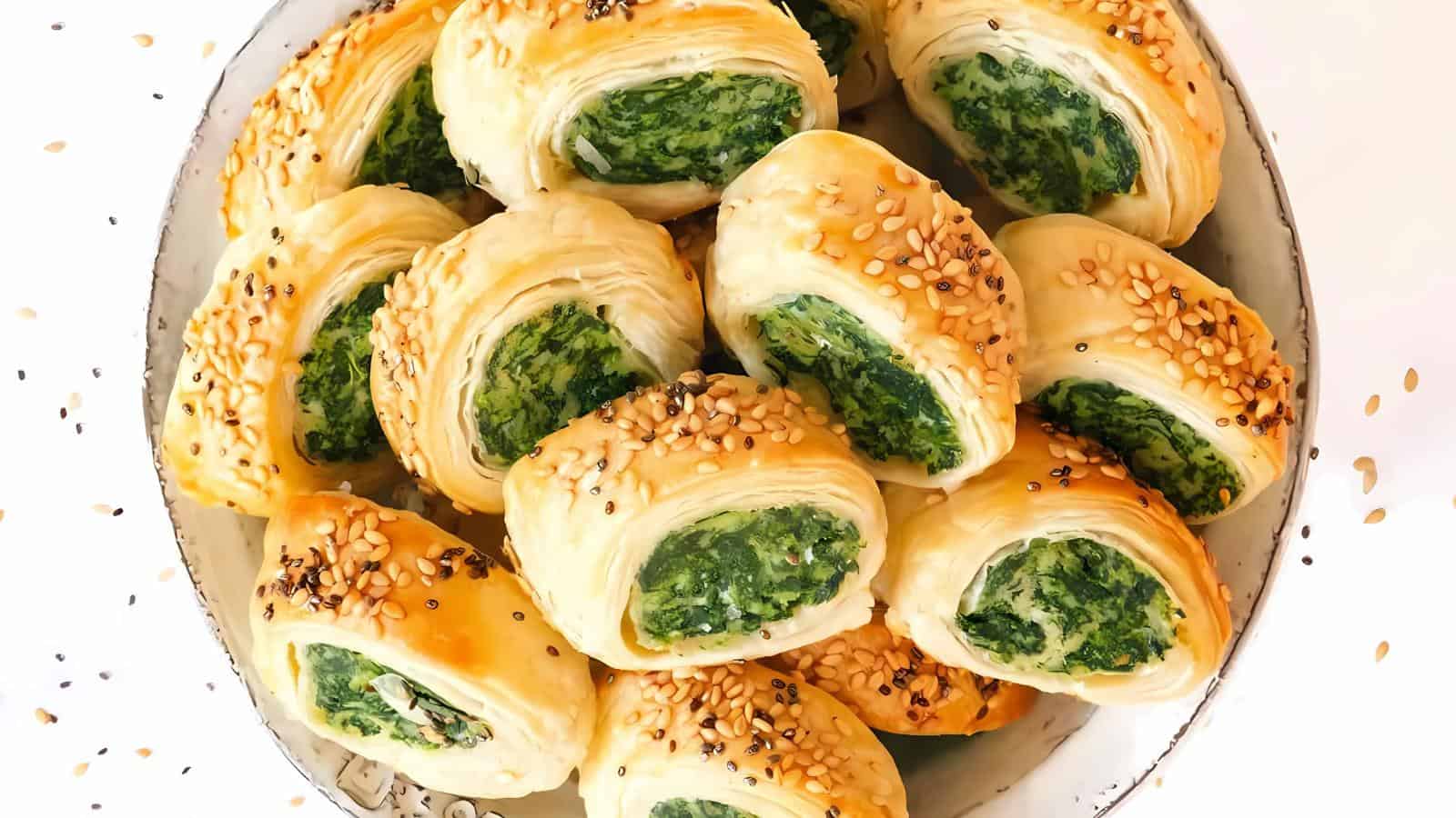 A bowl filled with golden puff pastry rolls stuffed with spinach filling and topped with sesame seeds.