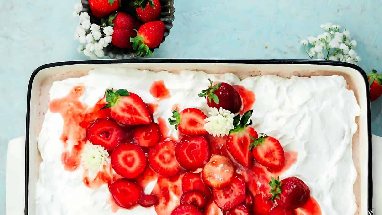 A rectangular dish with a dessert topped with whipped cream, sliced strawberries, and white flowers, next to a small bowl of whole strawberries and flowers.