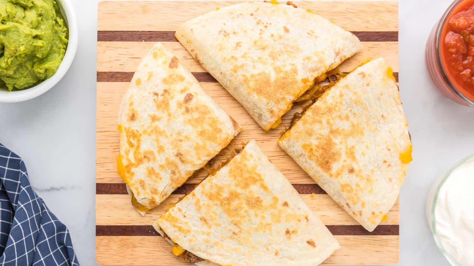 Four cheese quesadilla slices on a wooden cutting board, with bowls of guacamole, salsa, and sour cream nearby.