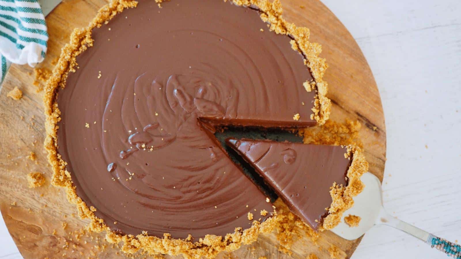 A chocolate tart with a graham cracker crust sits on a wooden board. One slice has been cut and is being lifted with a metal spatula.