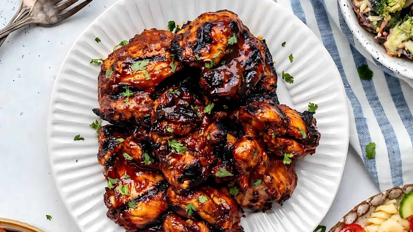 A plate of grilled barbecue chicken thighs garnished with chopped herbs, served on a white scalloped dish.