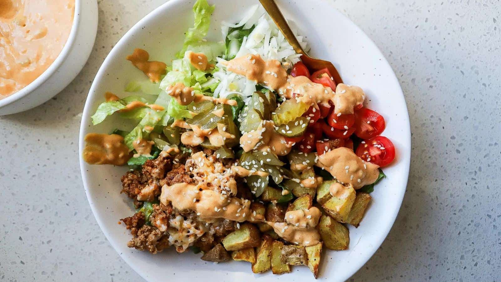 A bowl with ground beef, diced potatoes, lettuce, cherry tomatoes, pickles, shredded cheese, and sesame seeds, topped with a creamy sauce.