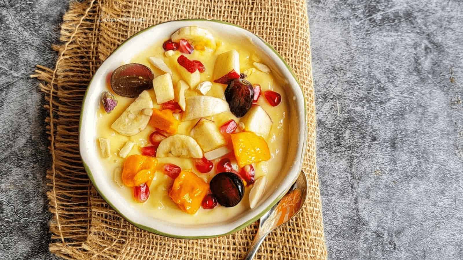 A bowl of fruit salad with yogurt, featuring banana, pomegranate seeds, grapes, papaya, and apple pieces on a burlap mat with a spoon beside it.