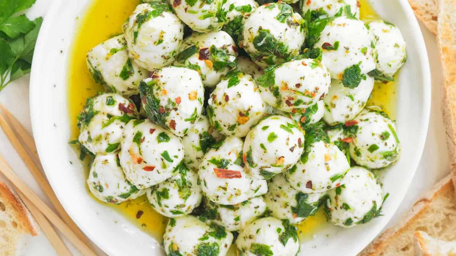 A bowl filled with marinated mozzarella balls topped with chopped herbs and chili flakes, surrounded by olive oil and slices of bread.