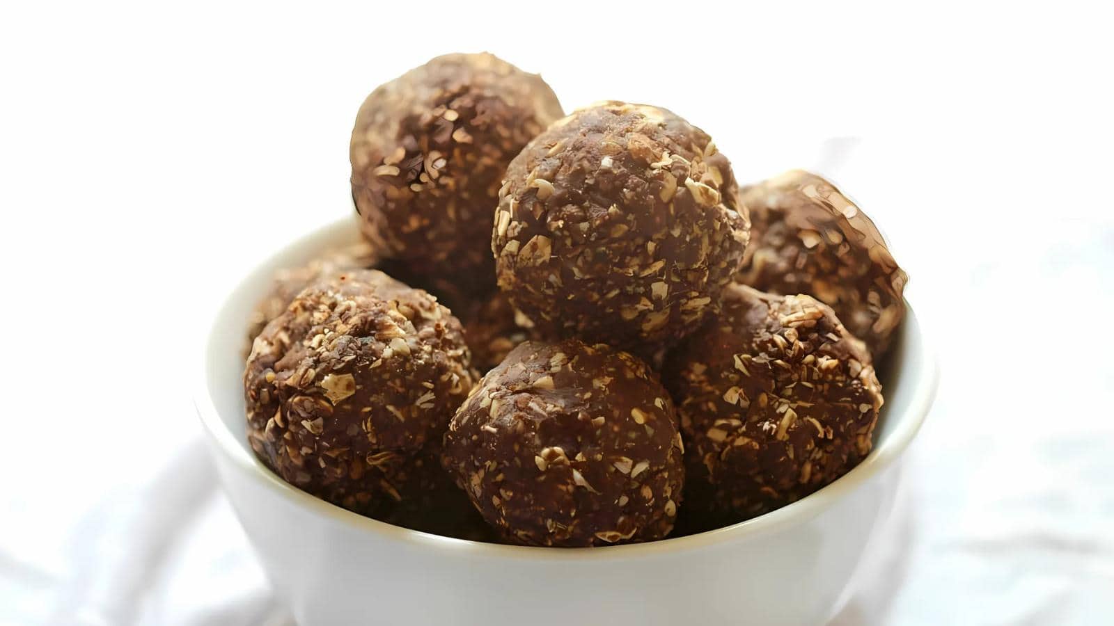 A white bowl filled with several chocolate and oat energy balls, placed on a white surface.