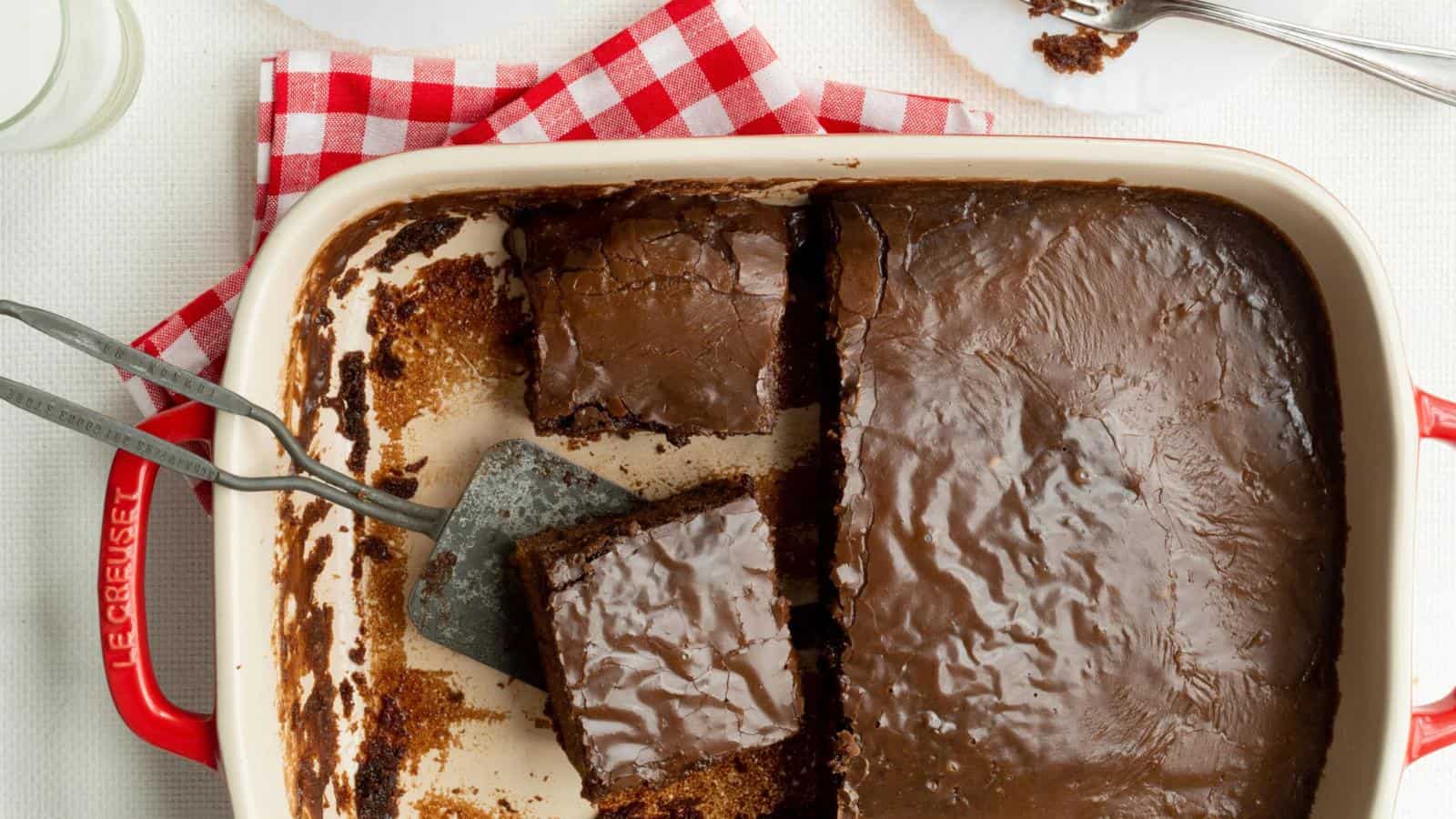 A pan of chocolate sheet cake with a few pieces cut out, a metal spatula, a red and white checked cloth, and a partially eaten piece on a plate nearby.