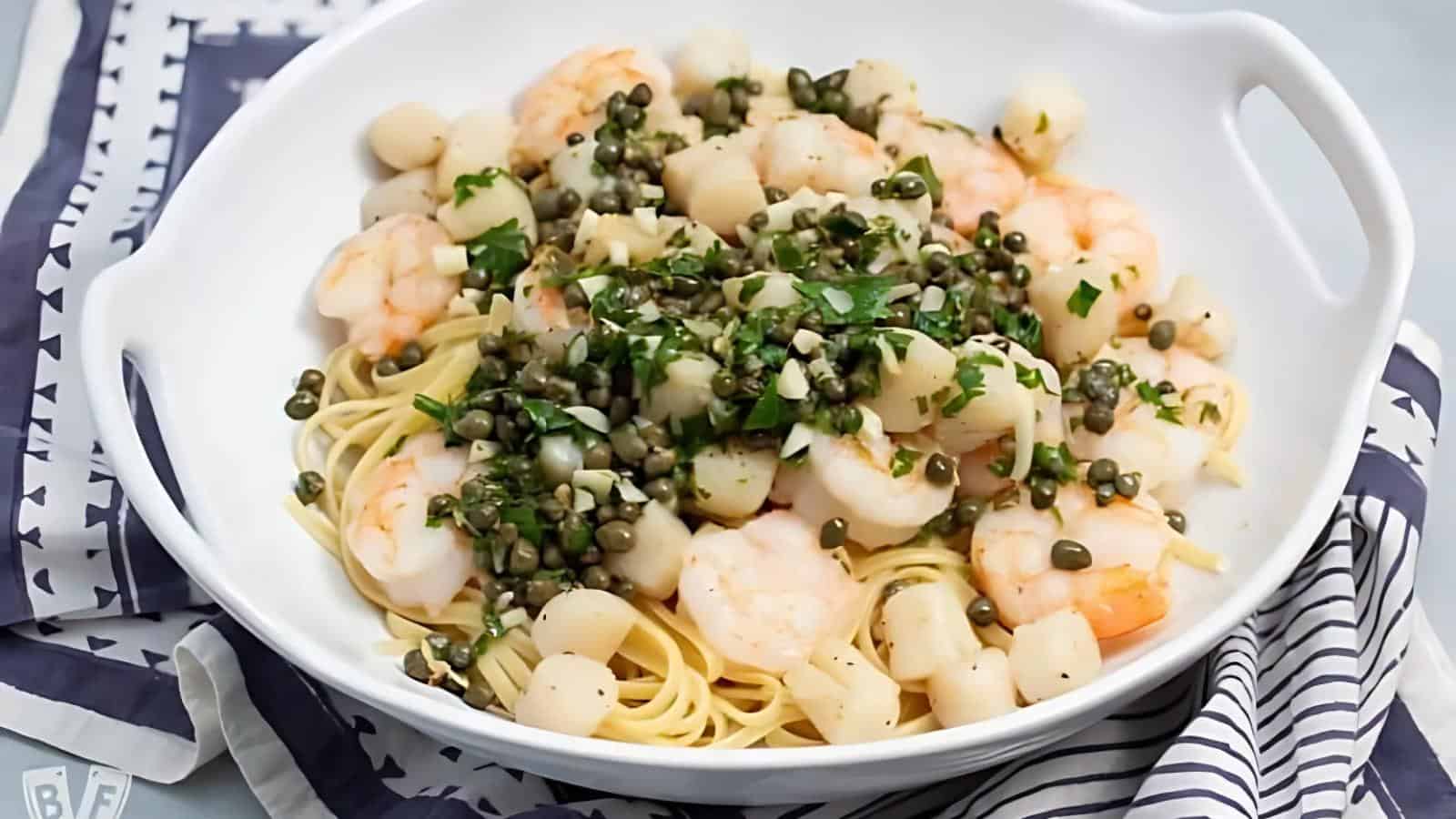 A white bowl filled with linguine, shrimp, scallops, capers, and chopped herbs sits on a patterned cloth.