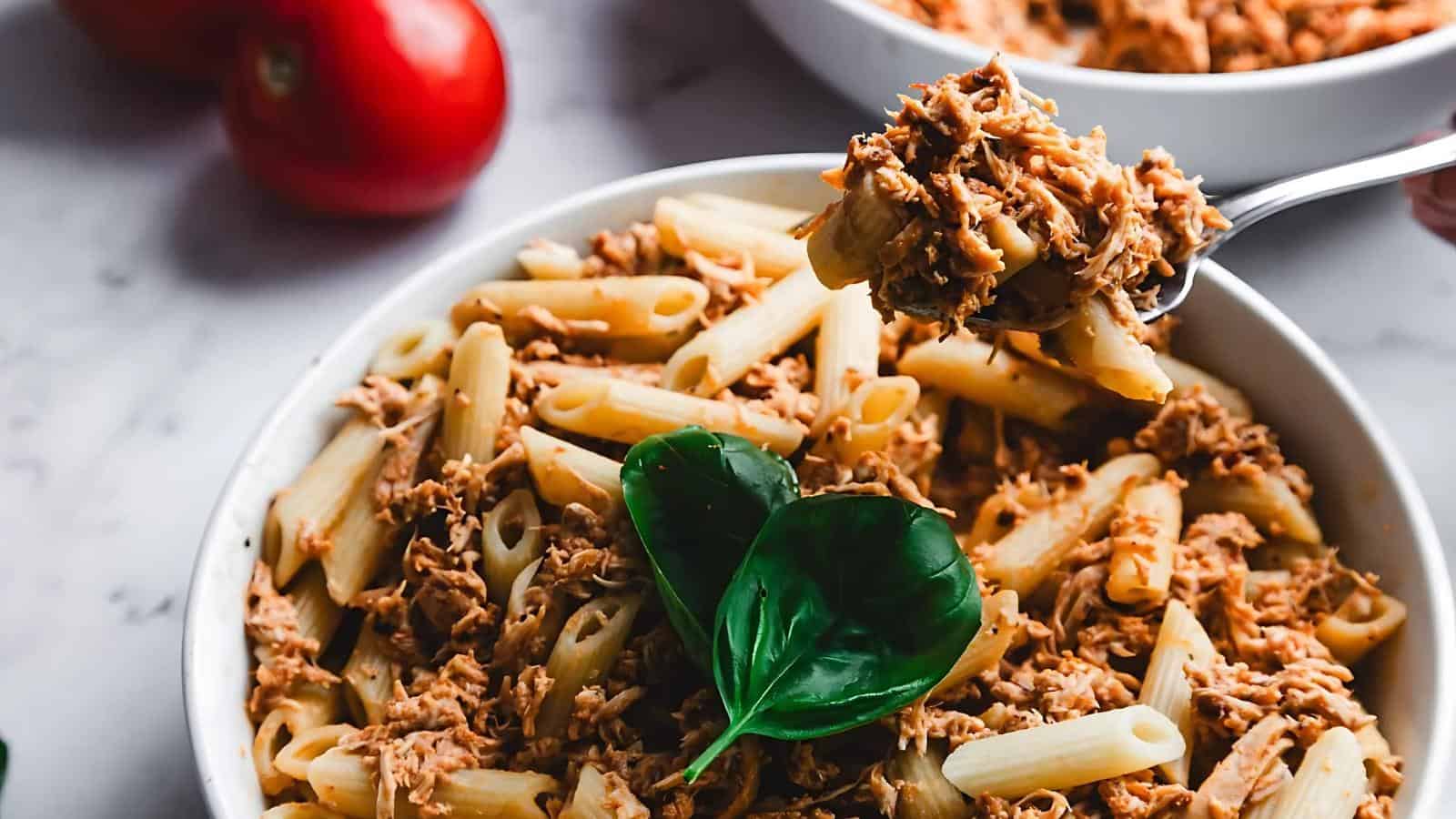 A bowl of penne pasta topped with shredded meat and garnished with fresh basil leaves, with a fork lifting a portion.