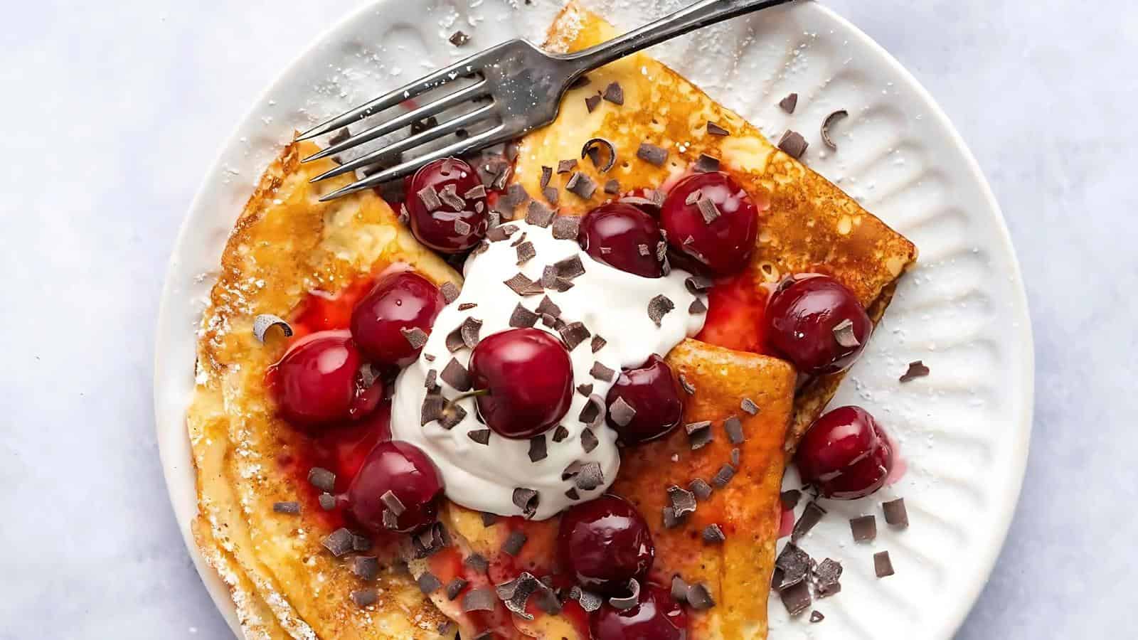 A plate with thin crepes topped with whipped cream, cherries, chocolate shavings, and a fork resting on the side.