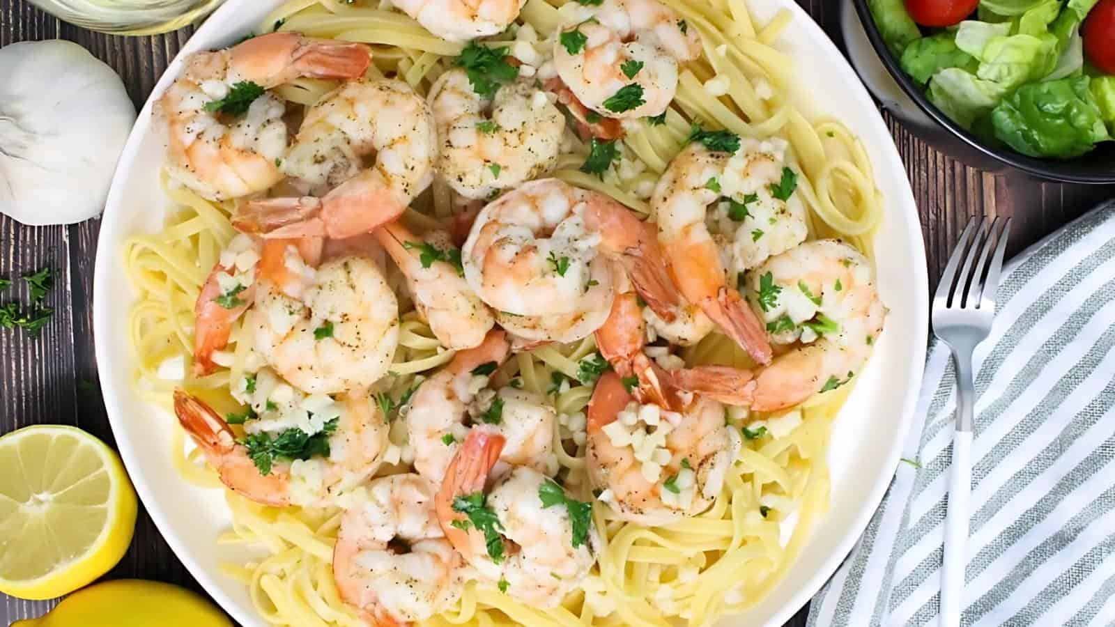 A plate of linguine pasta topped with cooked shrimp, garnished with chopped parsley and garlic, surrounded by salad, lemon slices, and a fork with a napkin.