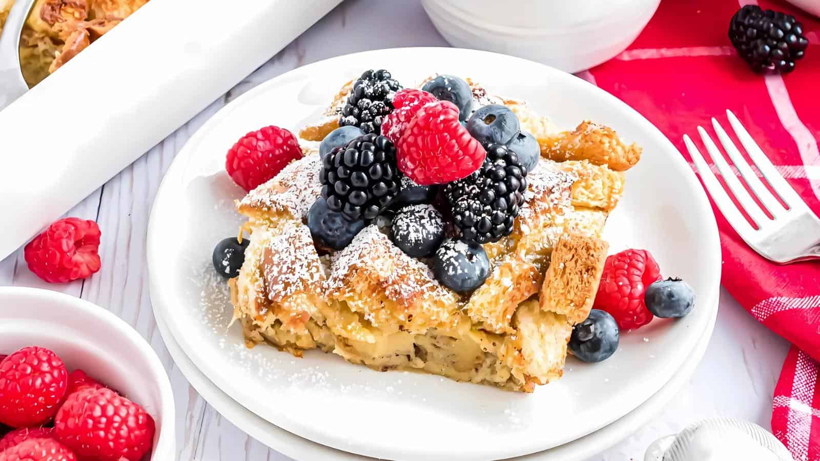 A slice of baked French toast topped with blackberries, blueberries, and raspberries, dusted with powdered sugar, on a white plate next to a red napkin.