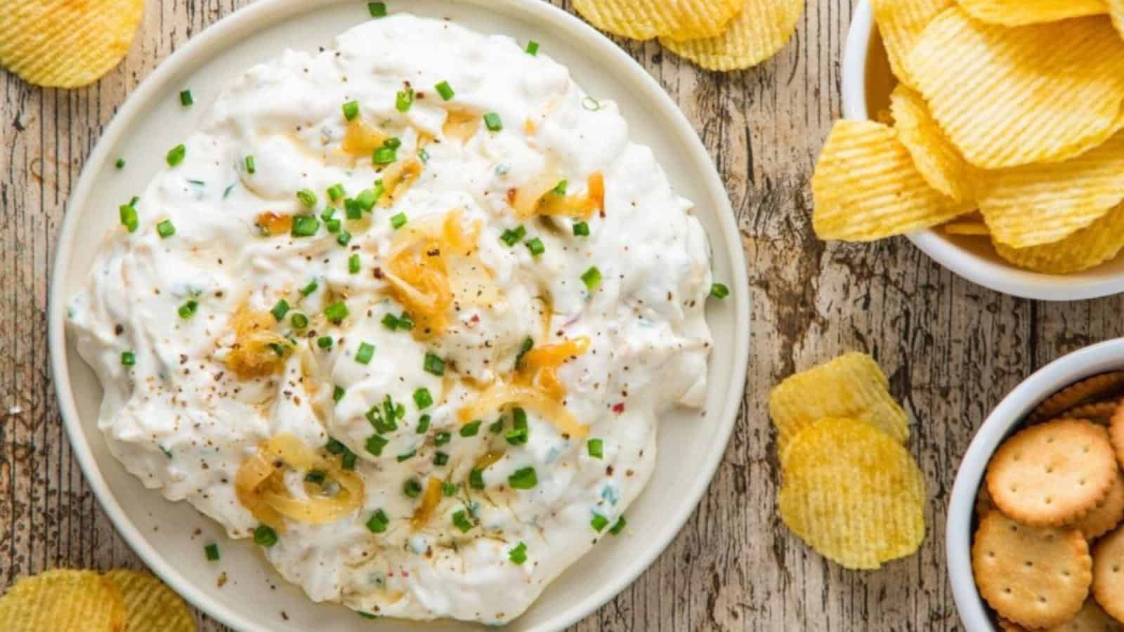 A bowl of creamy dip garnished with chopped chives and onions sits on a wooden table next to bowls of potato chips and round crackers.