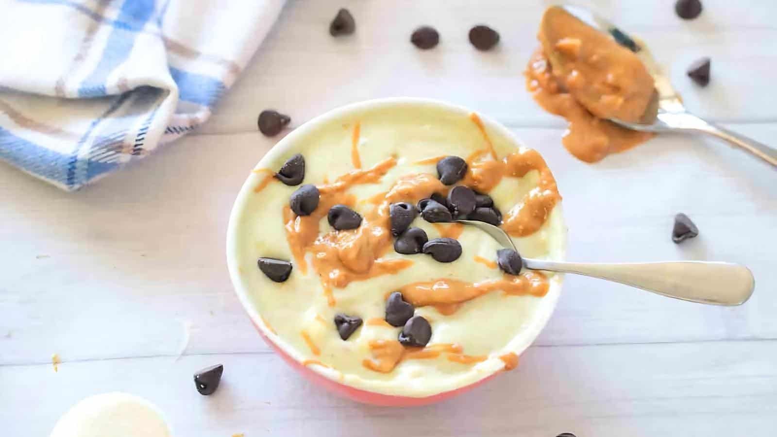 A bowl of yogurt topped with peanut butter and chocolate chips sits on a table with a spoon in it. A napkin and another spoon with peanut butter are nearby.