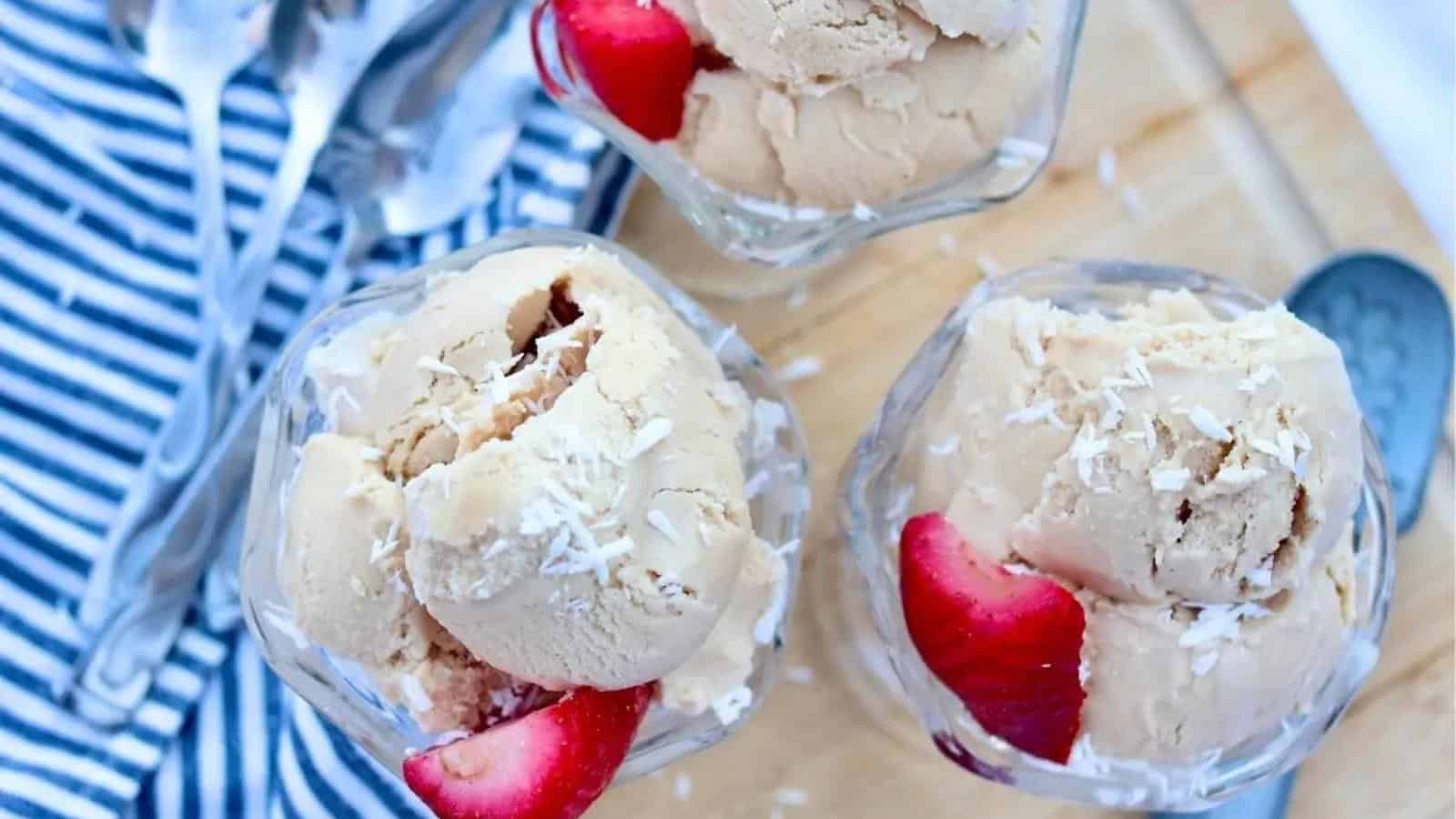 Three glass bowls of ice cream topped with shredded coconut and strawberry slices are placed on a wooden surface with a blue and white striped cloth and metal spoons nearby.