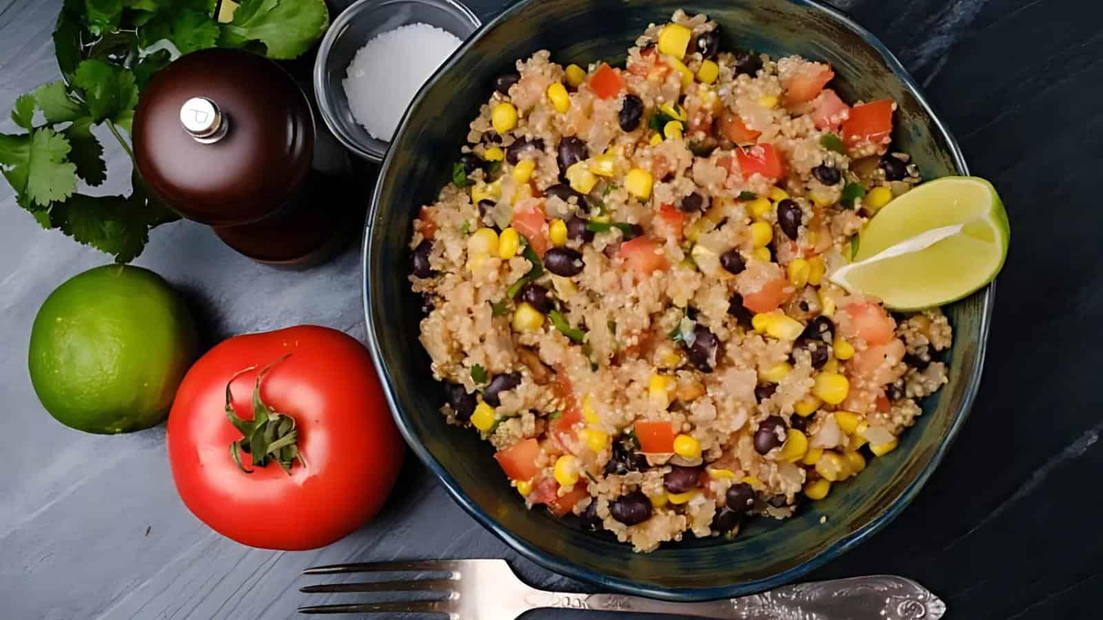 A bowl of quinoa salad with black beans, corn, tomatoes, and a lime wedge, surrounded by a tomato, lime, cilantro, salt, and pepper on a dark surface.