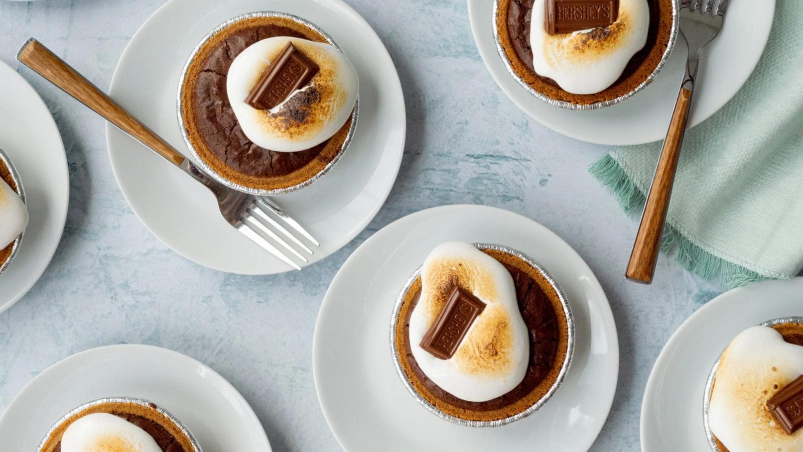 Mini chocolate pies topped with toasted marshmallows and Hershey’s chocolate squares are arranged on white plates with forks on a light surface.