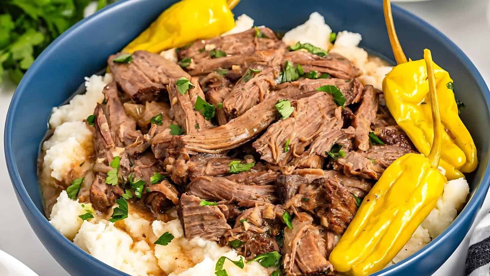 Shredded pot roast served over mashed potatoes, garnished with sliced parsley and whole pepperoncini peppers in a blue bowl.