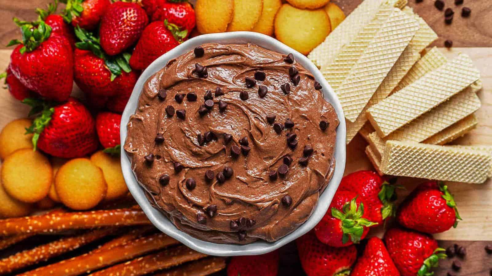 A bowl of chocolate dip with chocolate chips in the center, surrounded by strawberries, wafer cookies, vanilla wafers, and pretzel sticks on a wooden surface.
