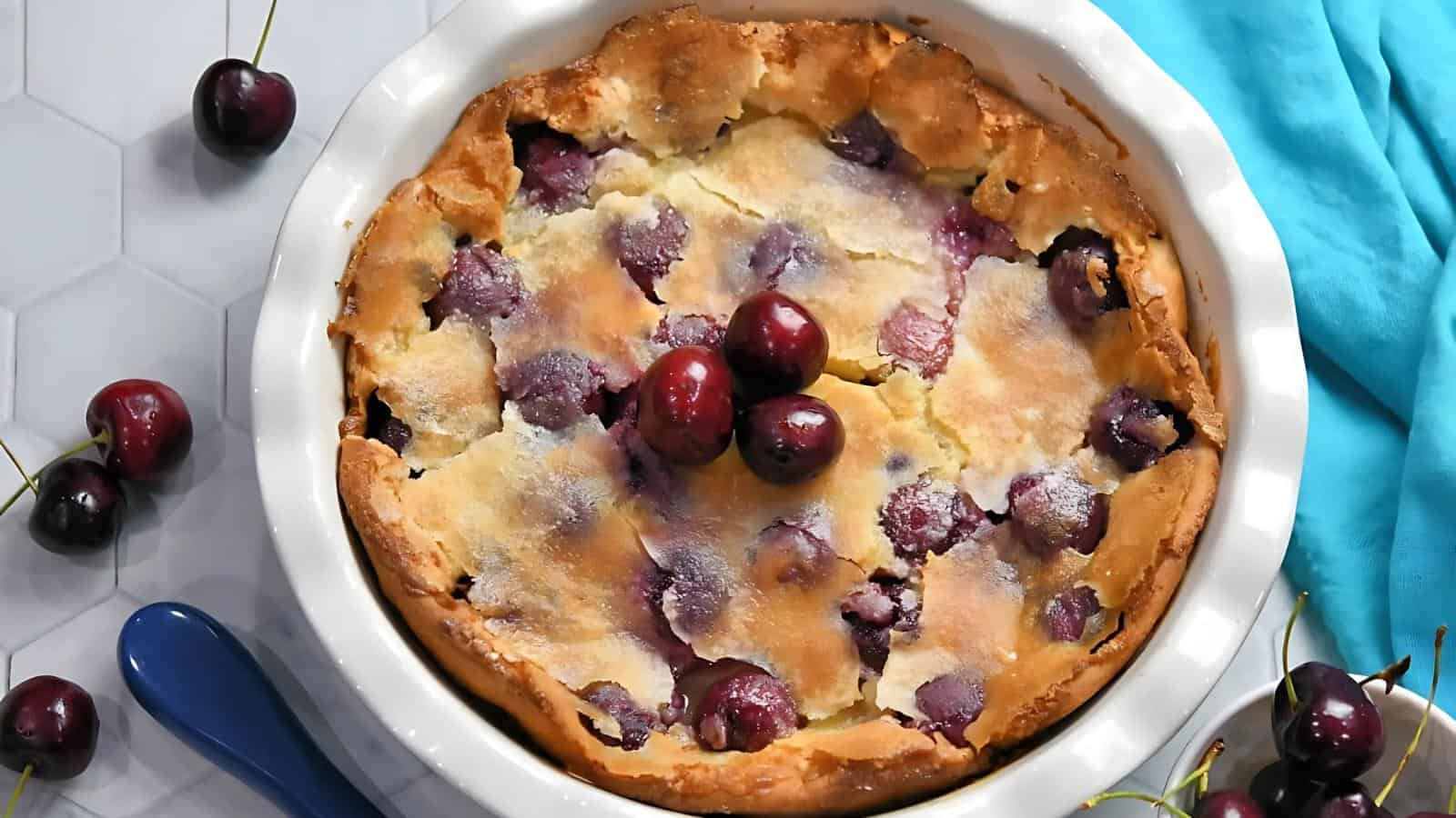 A baked cherry clafoutis in a round white dish, topped with whole cherries, surrounded by loose cherries and a blue napkin.