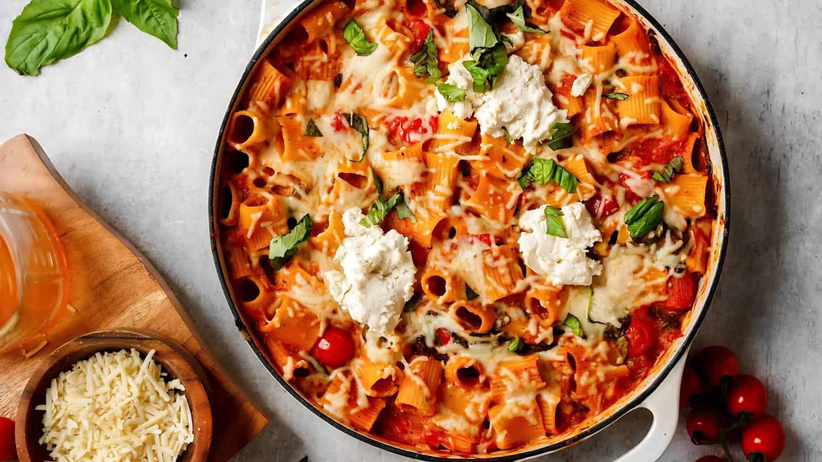 A baked pasta dish in a round pan topped with melted cheese, ricotta, and fresh basil, with a bowl of shredded cheese and a drink on the side.