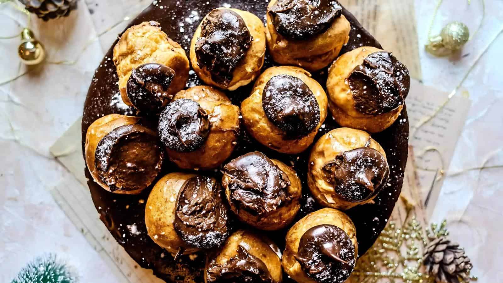 A close-up view of a plate filled with cream puffs topped with chocolate glaze and dusted with powdered sugar, seen from above.