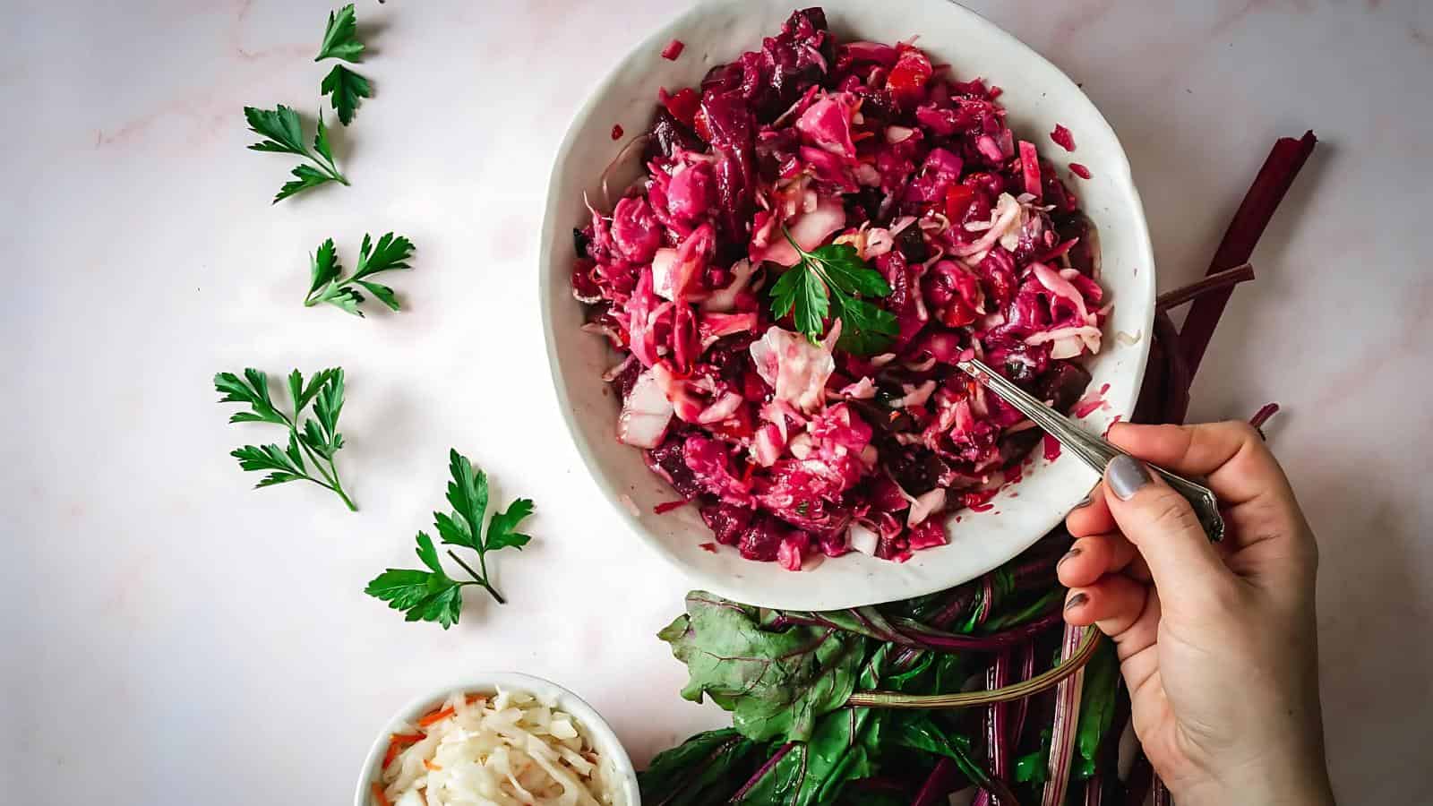 A hand holds a fork in a bowl of beet and cabbage salad, surrounded by fresh parsley and leafy greens on a white surface.
