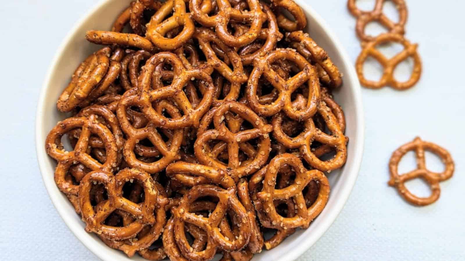 A white bowl filled with seasoned pretzel twists sits on a light surface, with a few pretzels scattered outside the bowl.