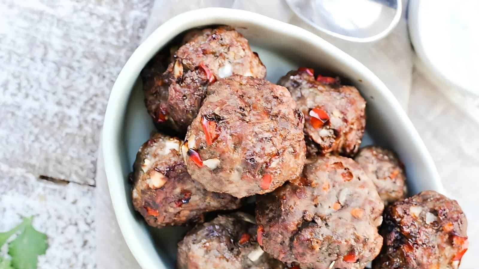 A bowl of baked meatballs with visible pieces of red pepper and herbs, placed on a light-colored surface.