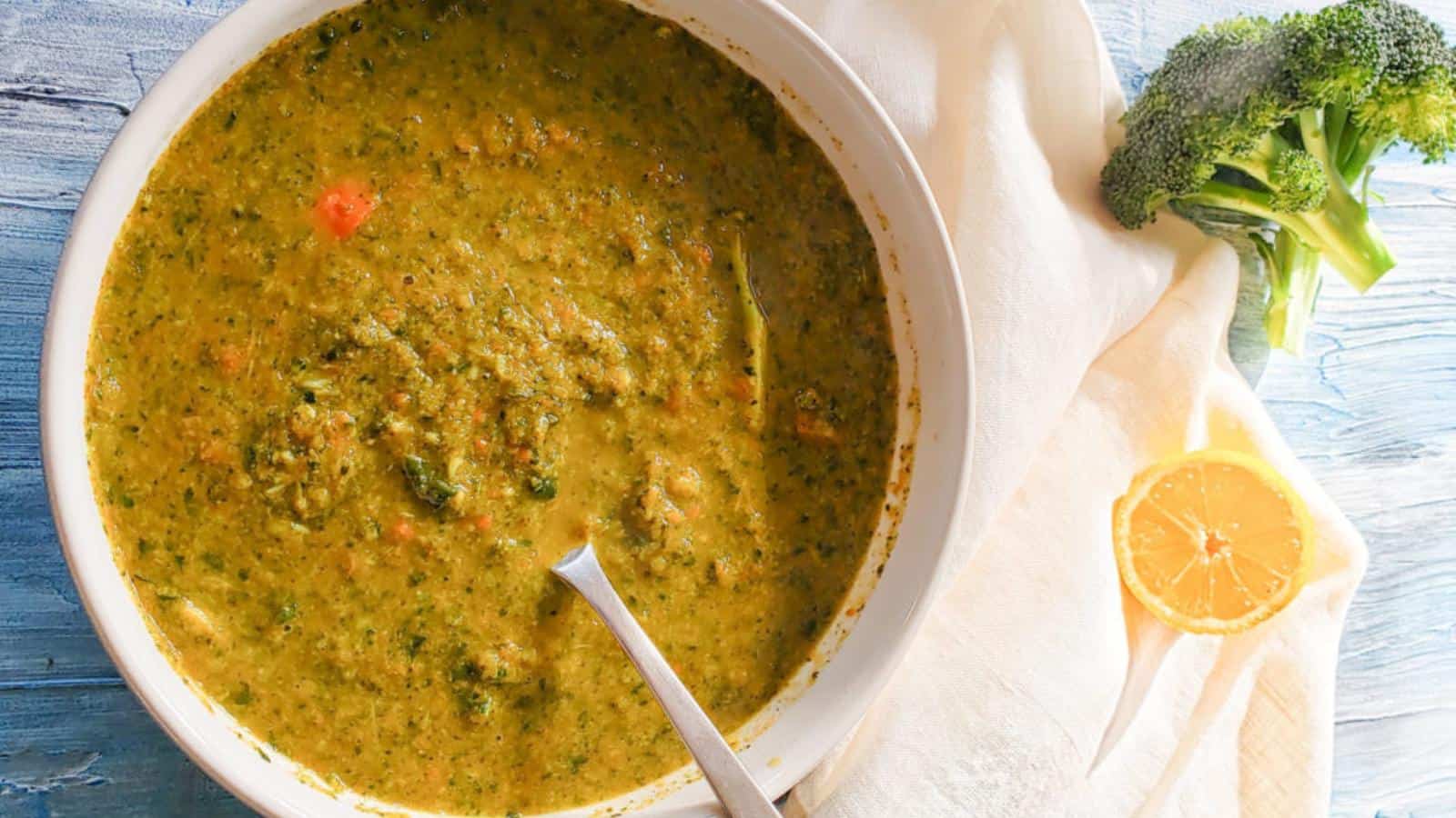 A bowl of thick vegetable soup with a spoon, placed on a white napkin next to a broccoli floret and a halved lemon.
