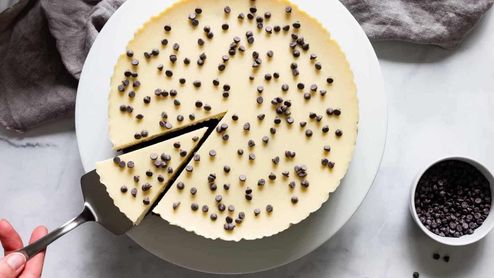A cheesecake topped with chocolate chips is being sliced, with one piece being lifted from the whole cake on a white plate next to a small bowl of chocolate chips.