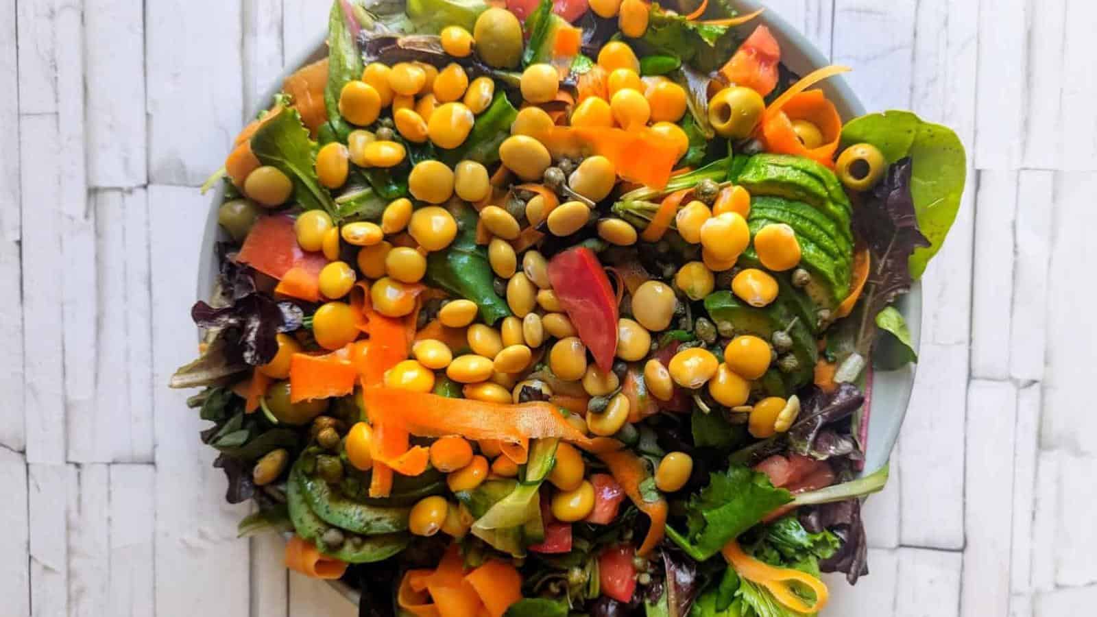 A bowl of mixed salad greens topped with sliced carrots, green olives, tomatoes, and yellow legumes on a white wooden surface.