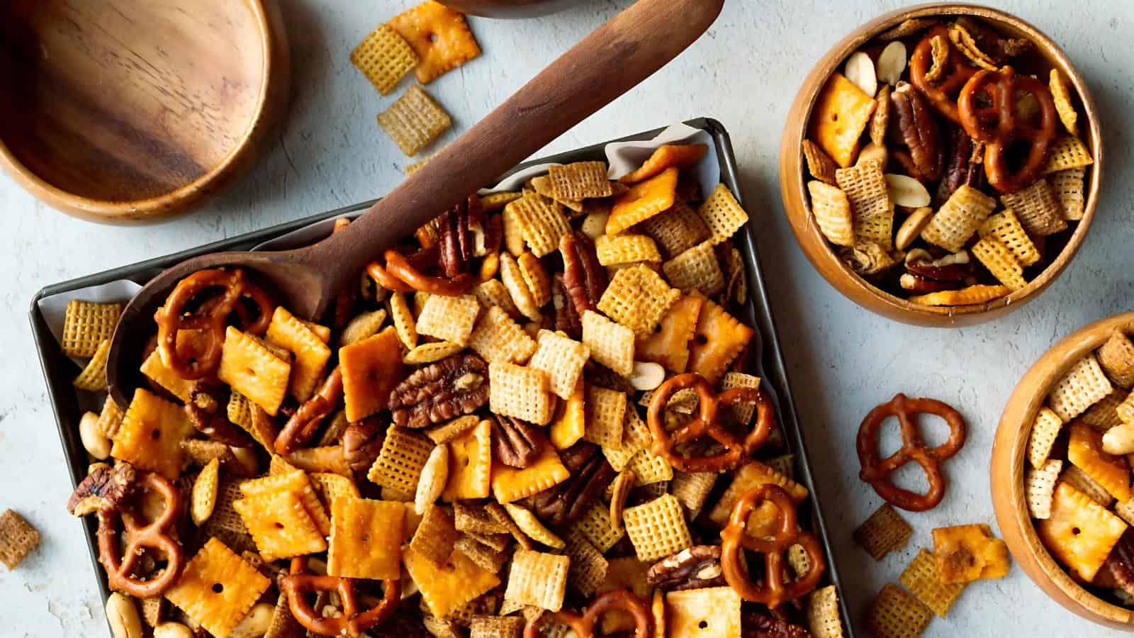A tray and bowls filled with a snack mix of pretzels, Chex cereal, cheese crackers, nuts, and seasoning, with a wooden spoon resting on the tray.