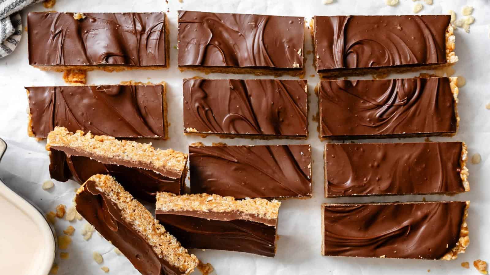 Twelve rectangular chocolate-topped bars with a crispy base are arranged on a sheet of parchment paper, with a few bars slightly overlapping.