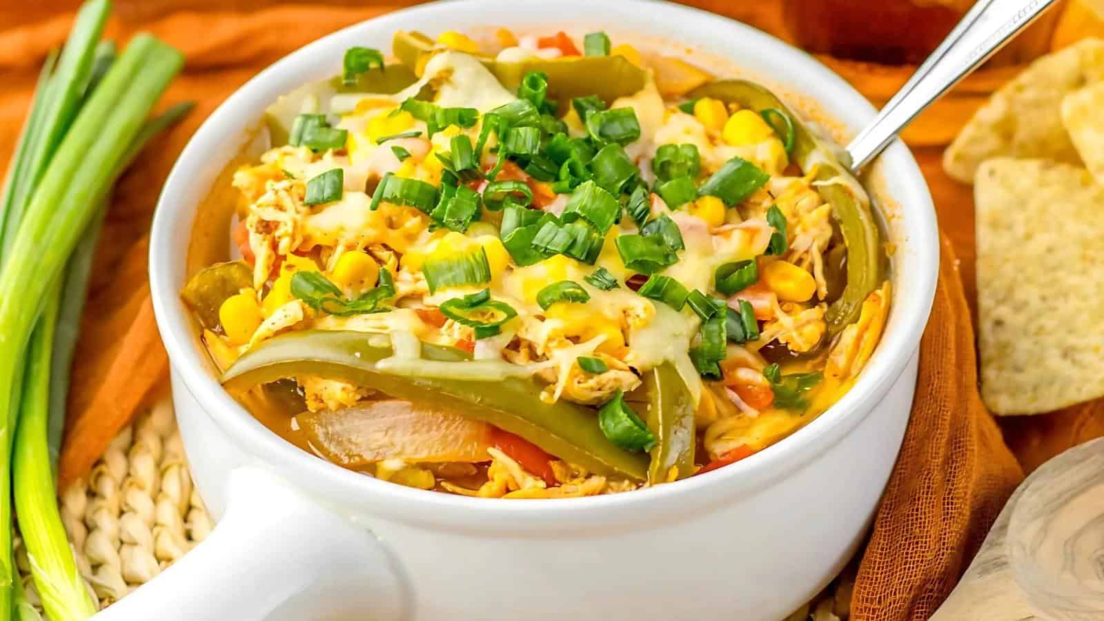 A bowl of chicken, cheese, green bell peppers, corn, and chopped green onions, served with a spoon and tortilla chips on the side.