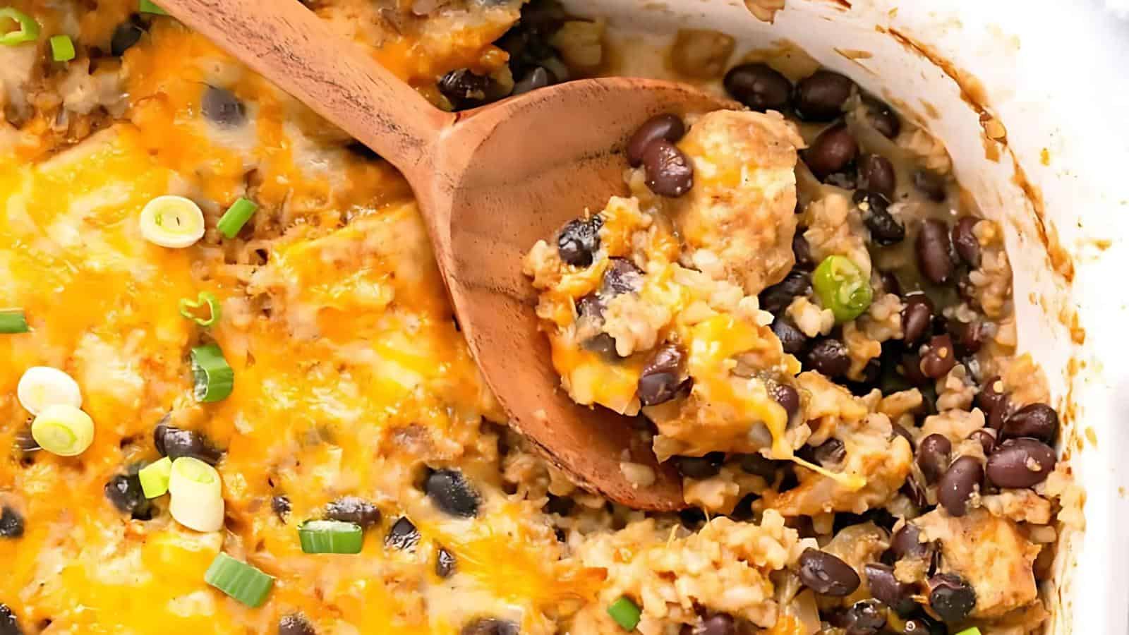 A wooden spoon scoops a cheesy casserole containing black beans, rice, green onions, and melted cheese from a baking dish.