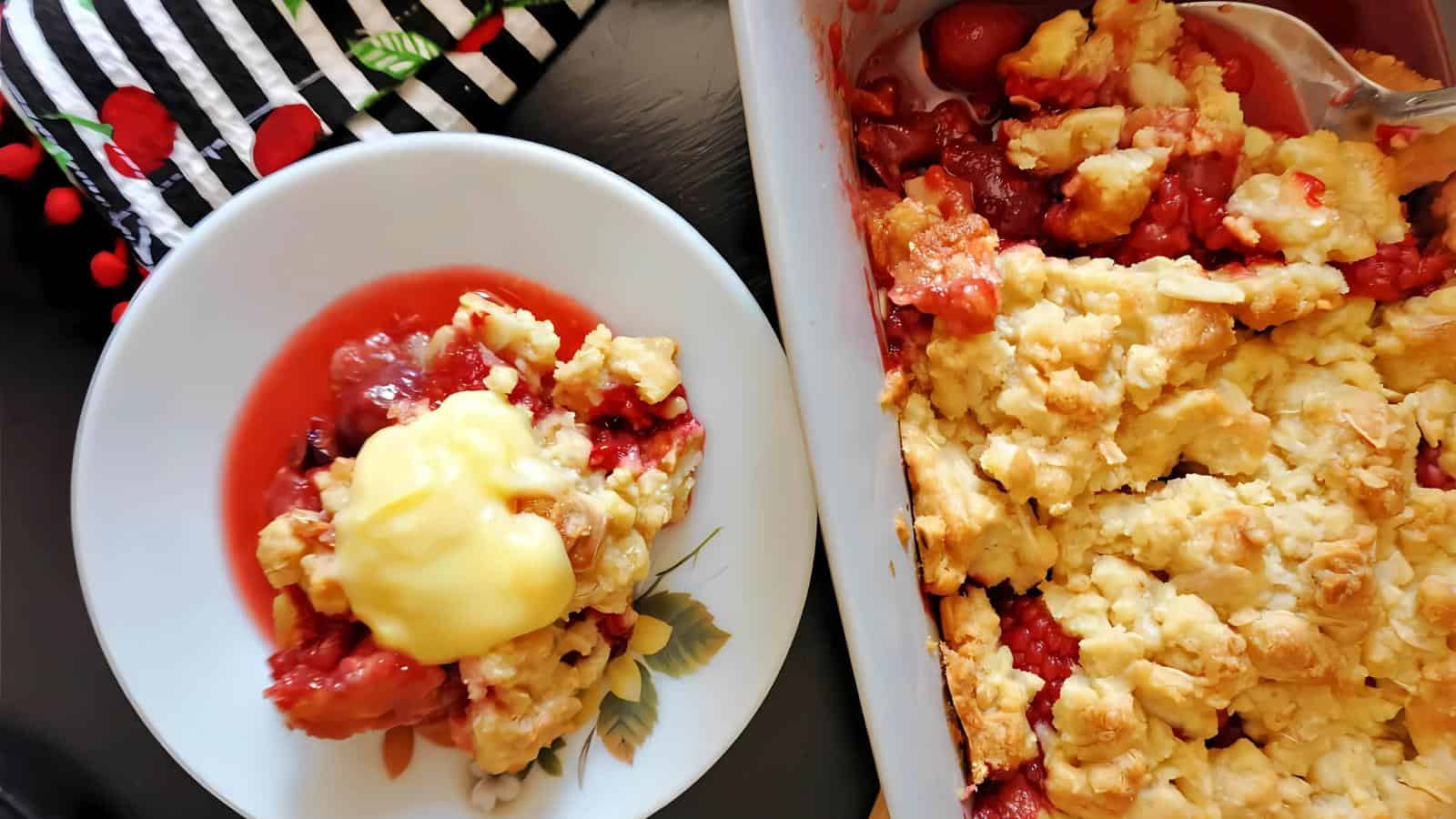 A serving of fruit cobbler with custard on a plate next to a baking dish of cobbler, placed on a table beside a striped cloth with red cherry patterns.