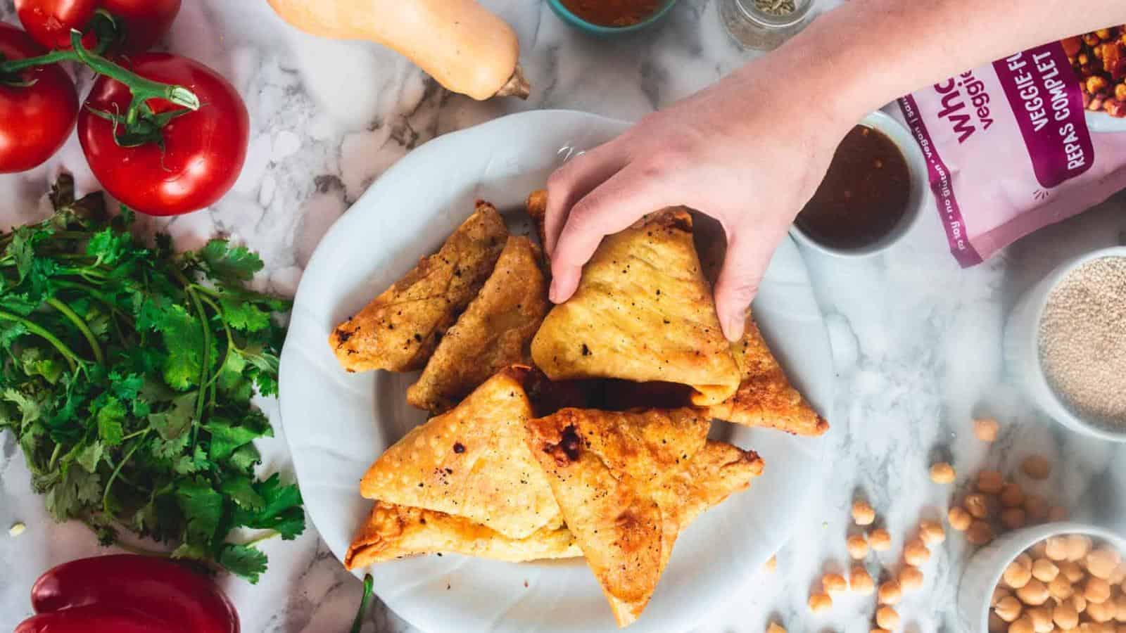 A hand reaches for a triangular pastry on a plate, surrounded by fresh tomatoes, cilantro, chickpeas, a butternut squash, and other cooking ingredients on a marble surface.