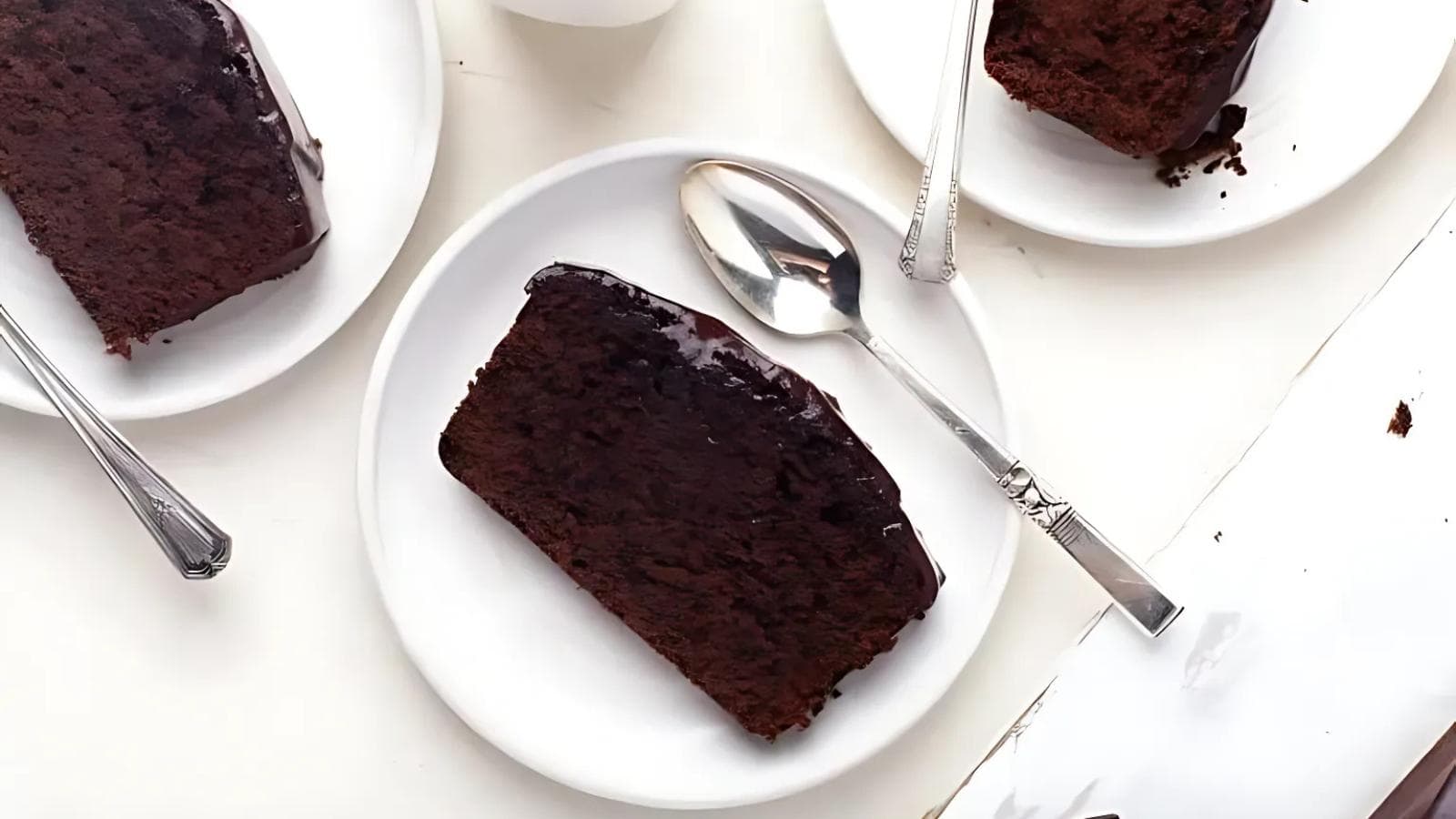 A slice of chocolate cake with a glossy chocolate topping on a white plate with a spoon, alongside other cake slices on similar plates.