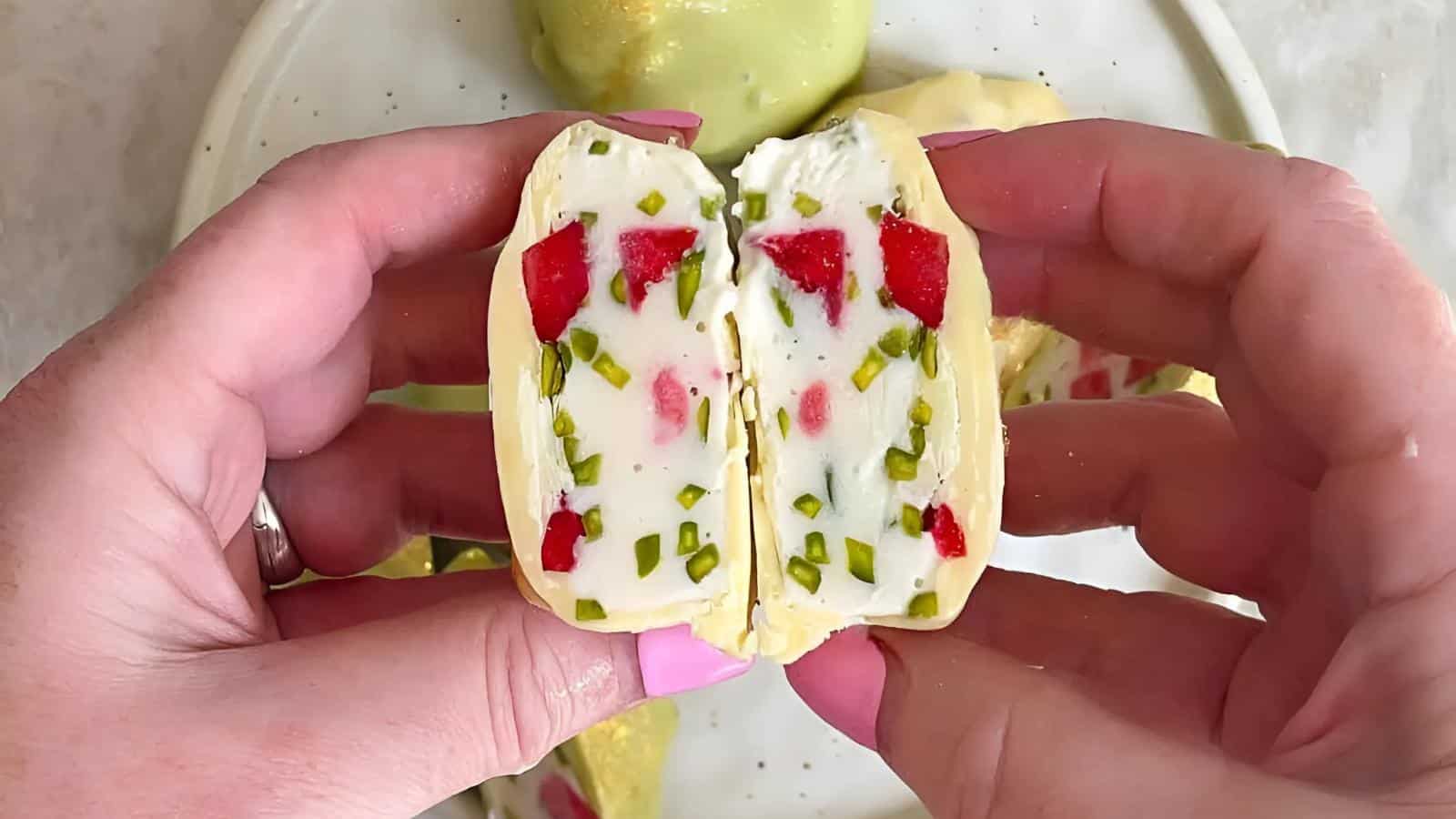 A person holds a halved dessert with a white creamy filling containing red and green pieces, coated in a yellow outer layer.