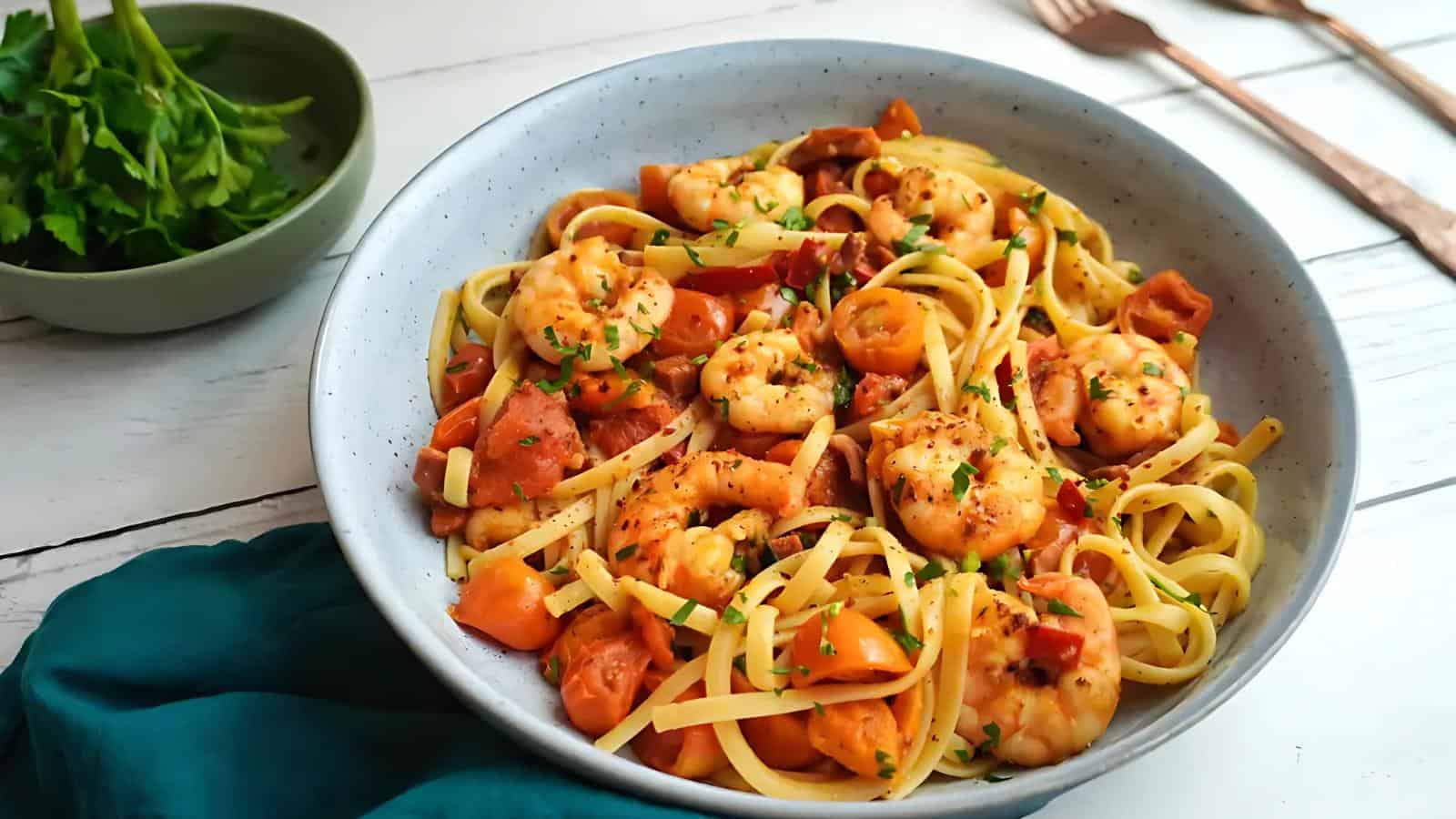 A bowl of linguine pasta topped with shrimp, chopped tomatoes, and herbs sits on a white table next to a bowl of fresh parsley.