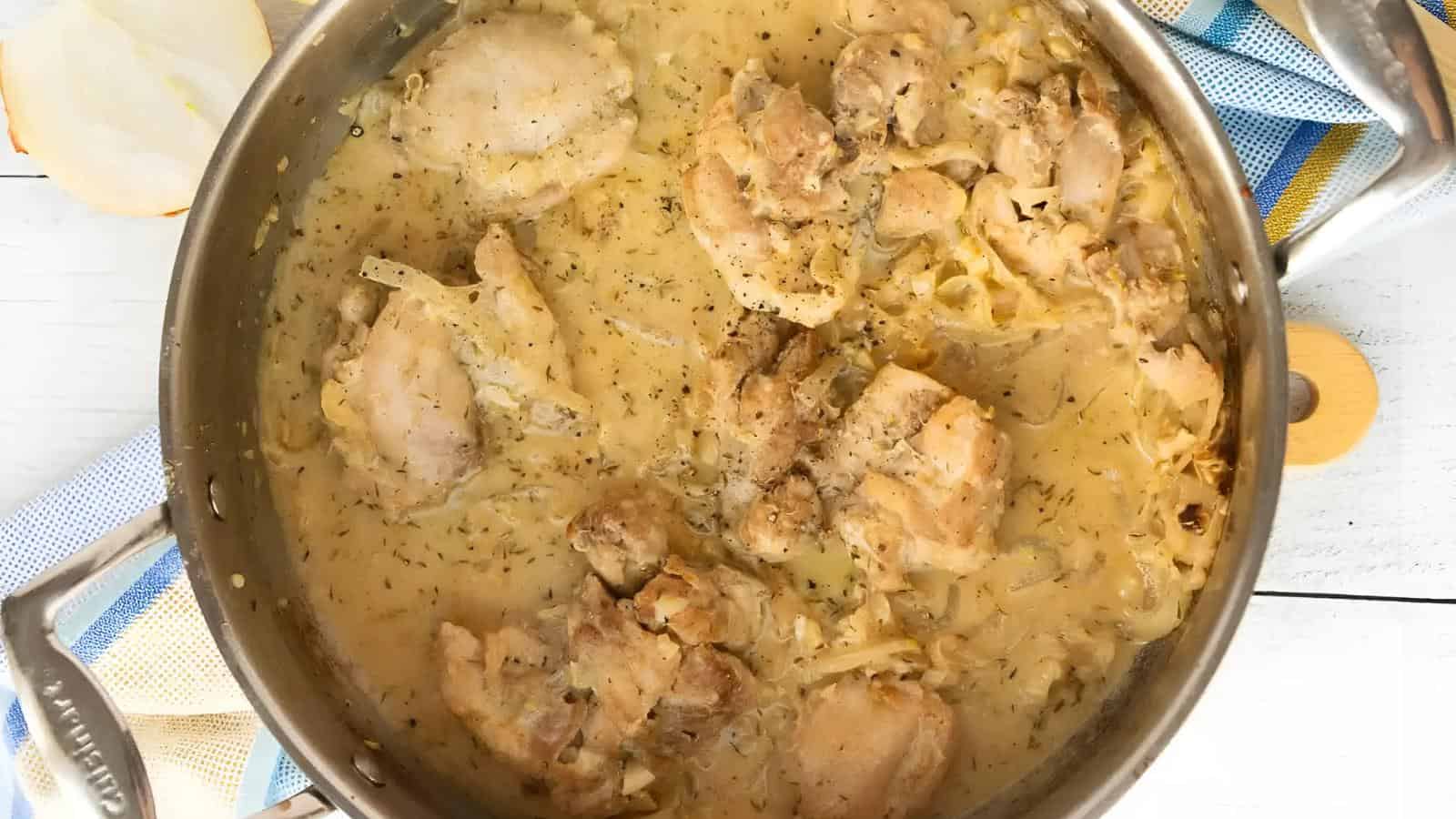 Chicken pieces simmer in a creamy sauce with onions and seasonings in a stainless steel pan, viewed from above.