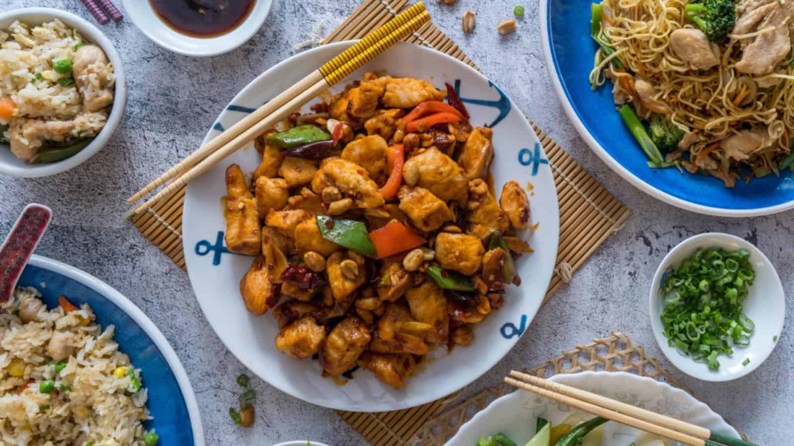 A plate of stir-fried chicken with vegetables and peanuts is surrounded by bowls of fried rice, noodles, sauce, and chopped green onions on a table.