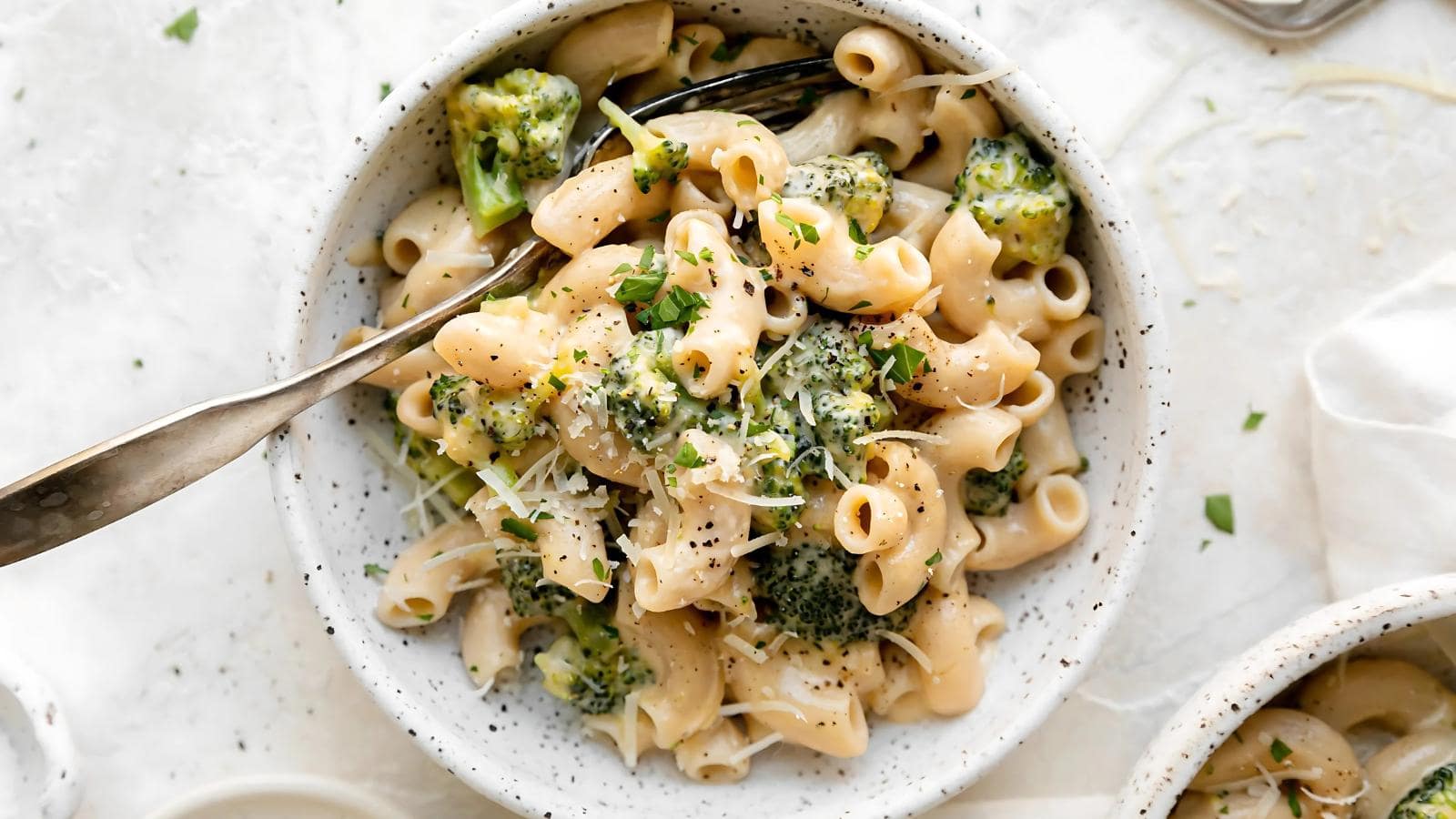 A bowl of macaroni pasta mixed with broccoli, topped with grated cheese and black pepper, with a fork resting inside.