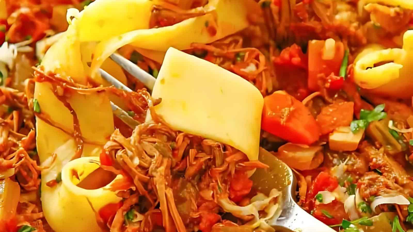 A close-up of pappardelle pasta mixed with shredded meat, diced tomatoes, and herbs, with a fork lifting a portion from the plate.