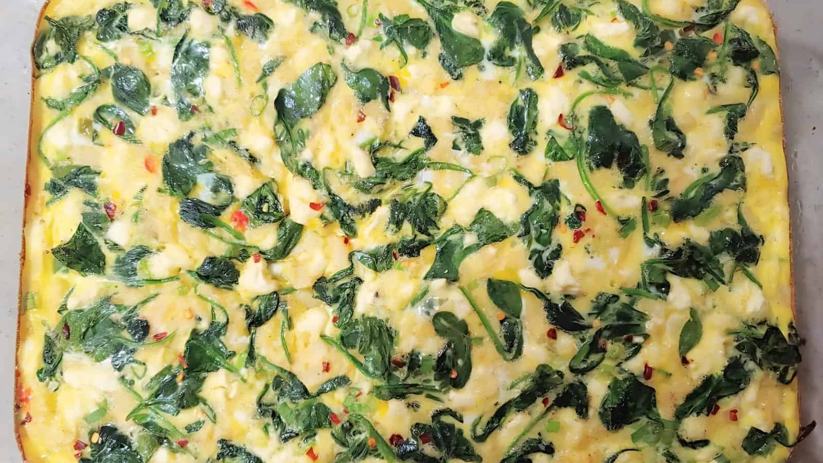 A baked egg casserole with visible spinach leaves and red pepper flakes spread evenly on the surface.
