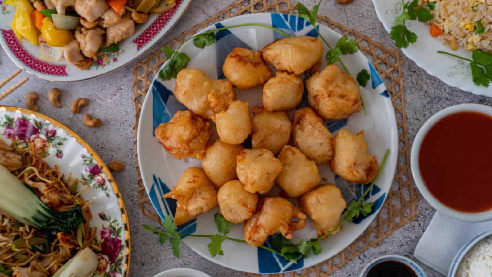 A plate of golden fried battered chicken pieces is surrounded by various side dishes, including stir-fried vegetables, fried rice, a bowl of red sauce, and noodles.