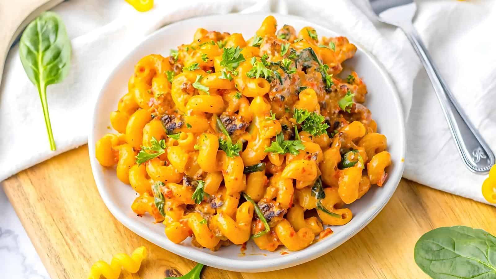 A plate of creamy macaroni pasta topped with chopped parsley, with spinach leaves and uncooked pasta scattered on a wooden surface nearby.