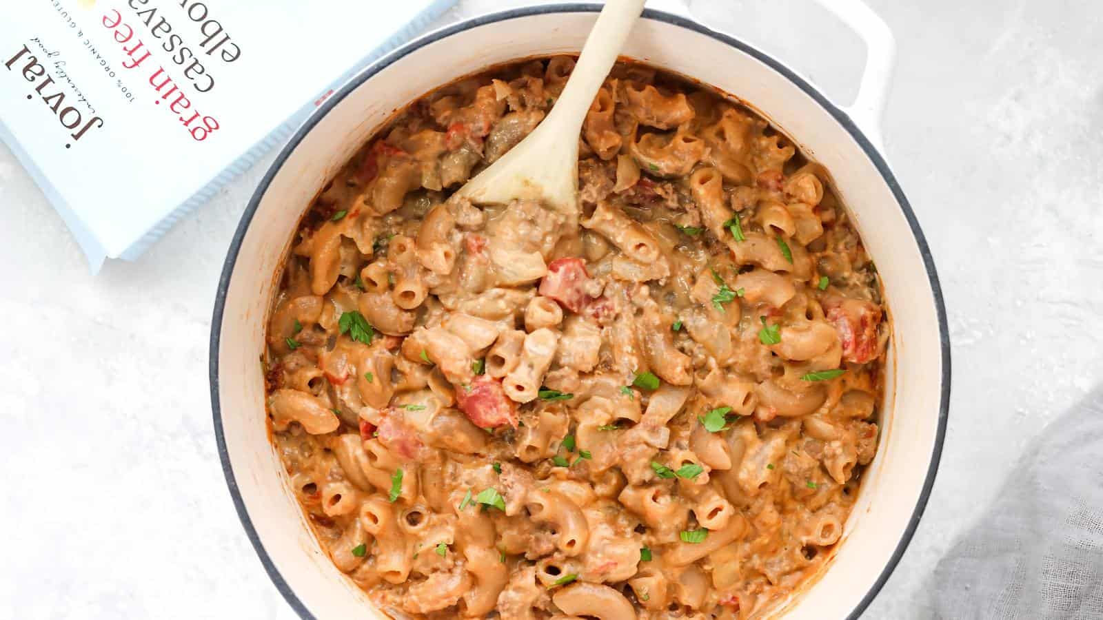 A white pot filled with creamy macaroni and ground beef casserole, garnished with parsley, sits beside an open cookbook on a light surface.
