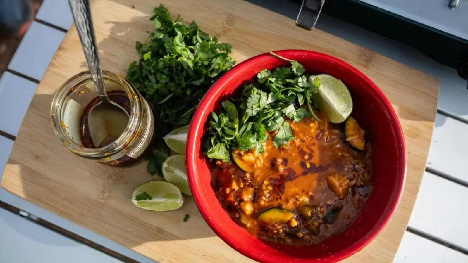 A red bowl of soup with vegetables and herbs sits on a wooden board alongside chopped cilantro, lime wedges, and a jar of pickled vegetables with a fork.